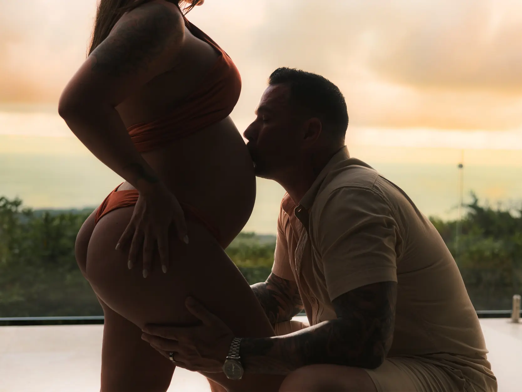 Luxury maternity photography at Kurá Boutique Hotel in Uvita. A tattooed partner kneels to kiss the expectant mother's bare belly during a beachside sunset session