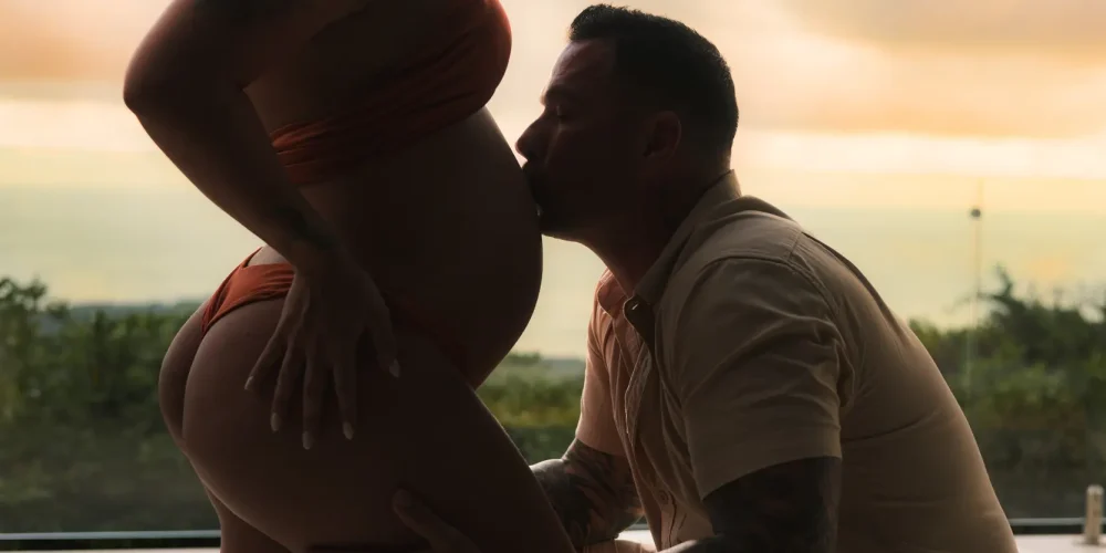 Luxury maternity photography at Kurá Boutique Hotel in Uvita. A tattooed partner kneels to kiss the expectant mother's bare belly during a beachside sunset session